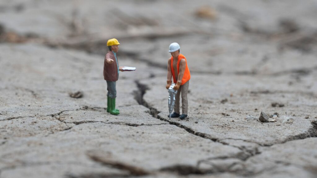 Miniature toy construction workers on cracked ground, one holds a clipboard, another a power tool | workforce strategy
