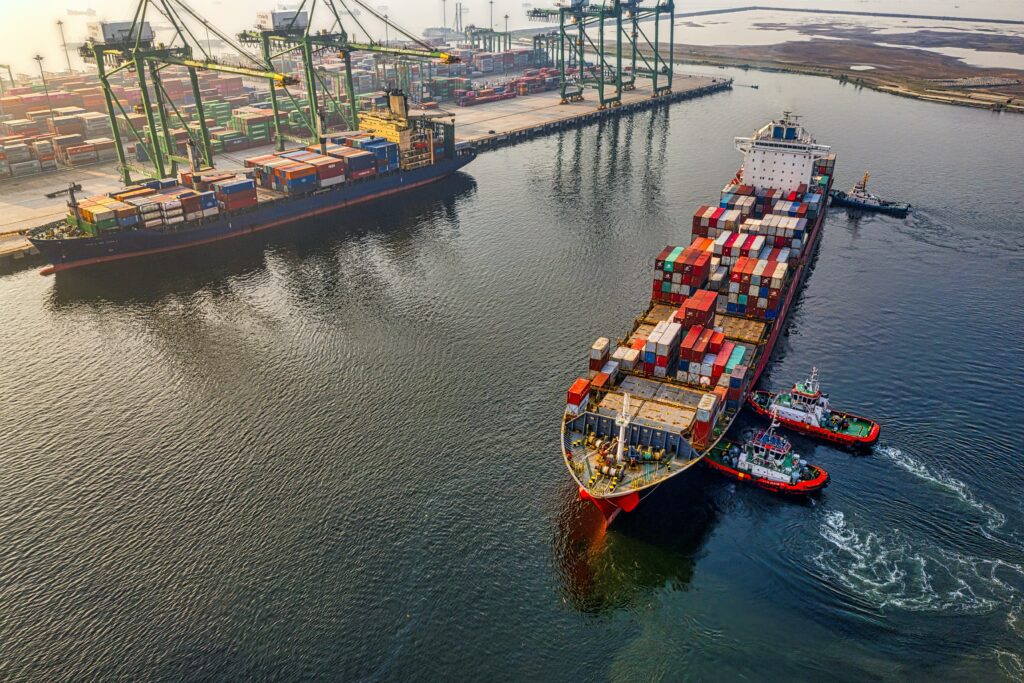 Overhead view of a cargo ship heavily laden wtih shipping containers being pushed into harbour with two tug boats | IFS supply chain management