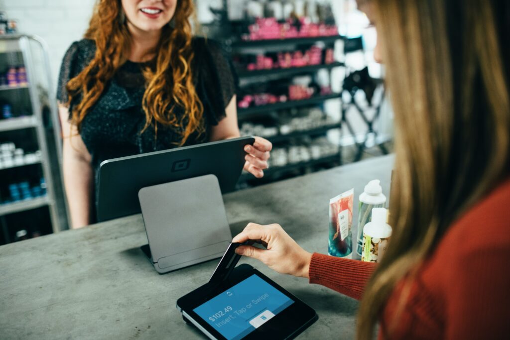 Two women standing at a shop counter, one of the women is purchasing items via a tablet and contactless payment | shop floor workers RamBase