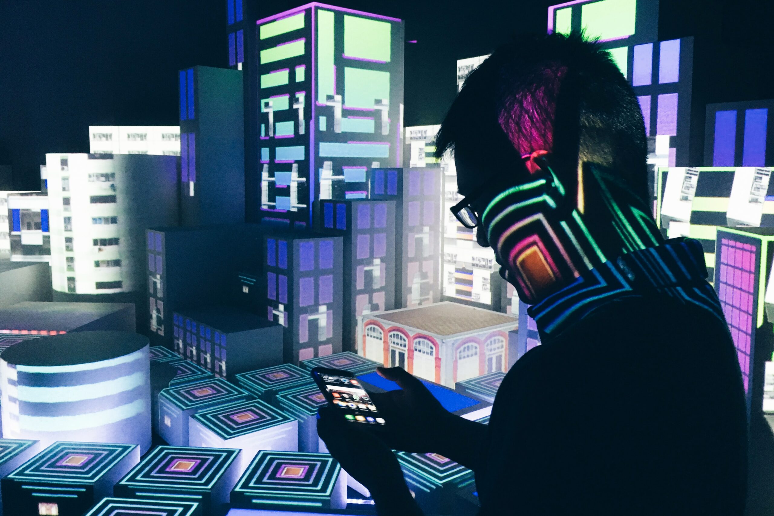 Abstract photo of a virtual city, with a silhouette of a man looking at his phone while standing over the neon-coloured cityscape | X4U Unit4