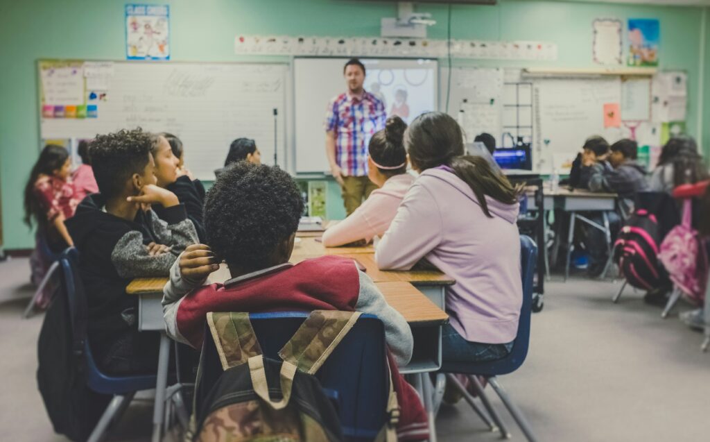 A classroom of middle-school children listening intently to a male teacher, standing at the front of the class | ServiceNow digital education