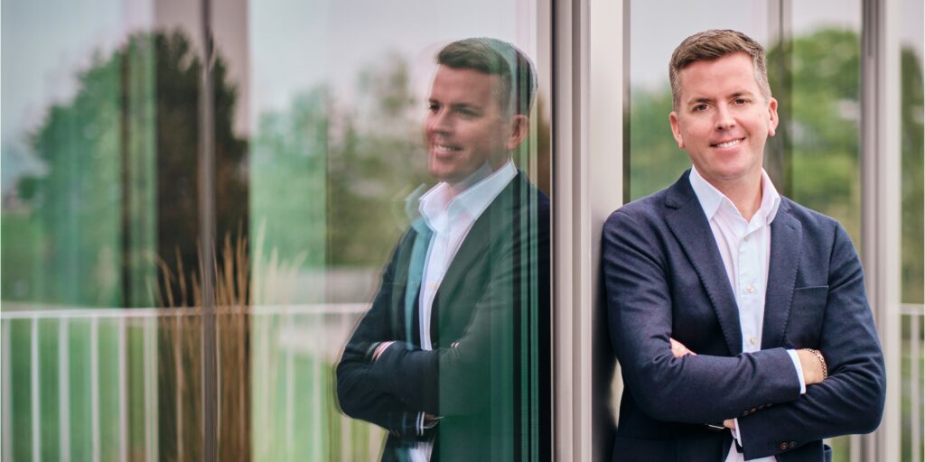 Image of Brian Duffy, CEO at SoftwareOne leaning against a window with greenery reflected | Photos by Kurt Rebry