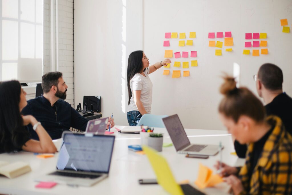 A woman presenting several postit notes on a wall to several people sitting at a meeting room desk | learning management GSS