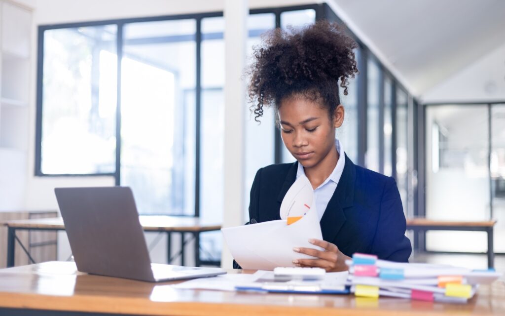 a woman in a white shirt and blue blazer sits at the work desk with a laptop looking through papers | Kyriba APIs finance