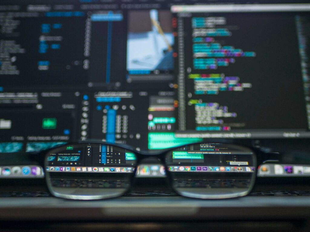 A blurred image of several computer screens chock full of code with a pair of glasses set on the desk in front of them - the angle shows through the glasses is in focus | third-party Rimini Street