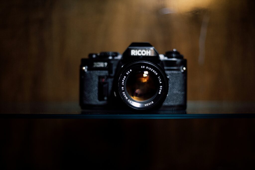 A moody coloured shot of a vintage Ricoh branded SLR camera against a dark, wooden background | Ricoh Company Rimini Street