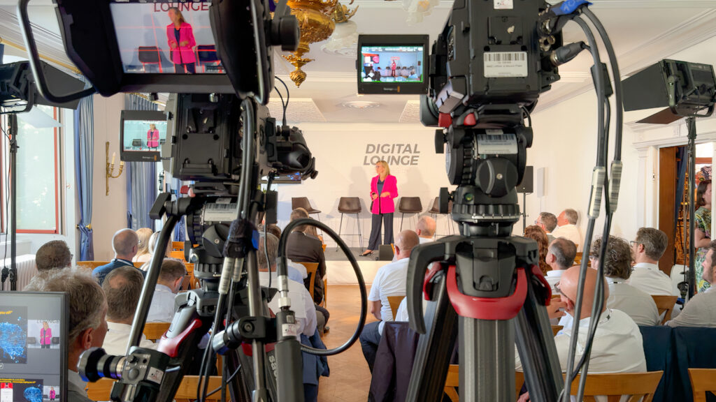 photo of a conference room with a woman in pink jacket on stage, two cameras filming her and audience sitting | Digital Lounge at Lakeside, Data Migration International (DMI), JiVS IMP, SAP