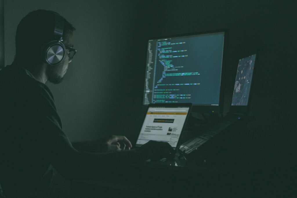 A darkened room with a man with big headphones sitting at 3 computer screens | Delinea