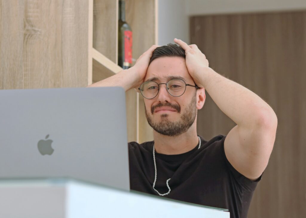 An exasperated man grasping his head in frustration while sitting in front of a macbook | Original Software Testing