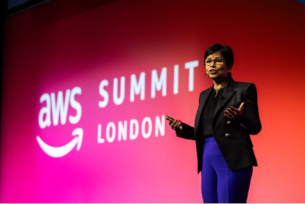 A speaker on stage at the AWS Summit in London, standing in front of a large screen with the event logo. The person is wearing a black blazer and blue pants, holding a presentation remote and speaking to the audience