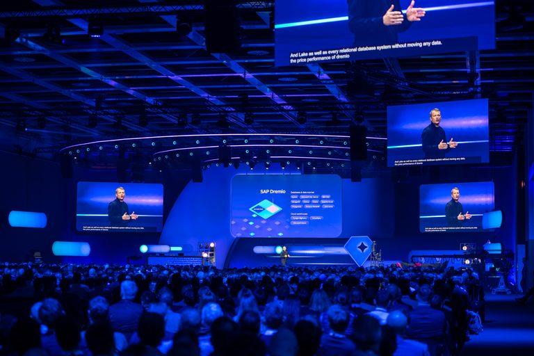 Conference speaker on stage in front of a large screen displaying the SAP logo and audience